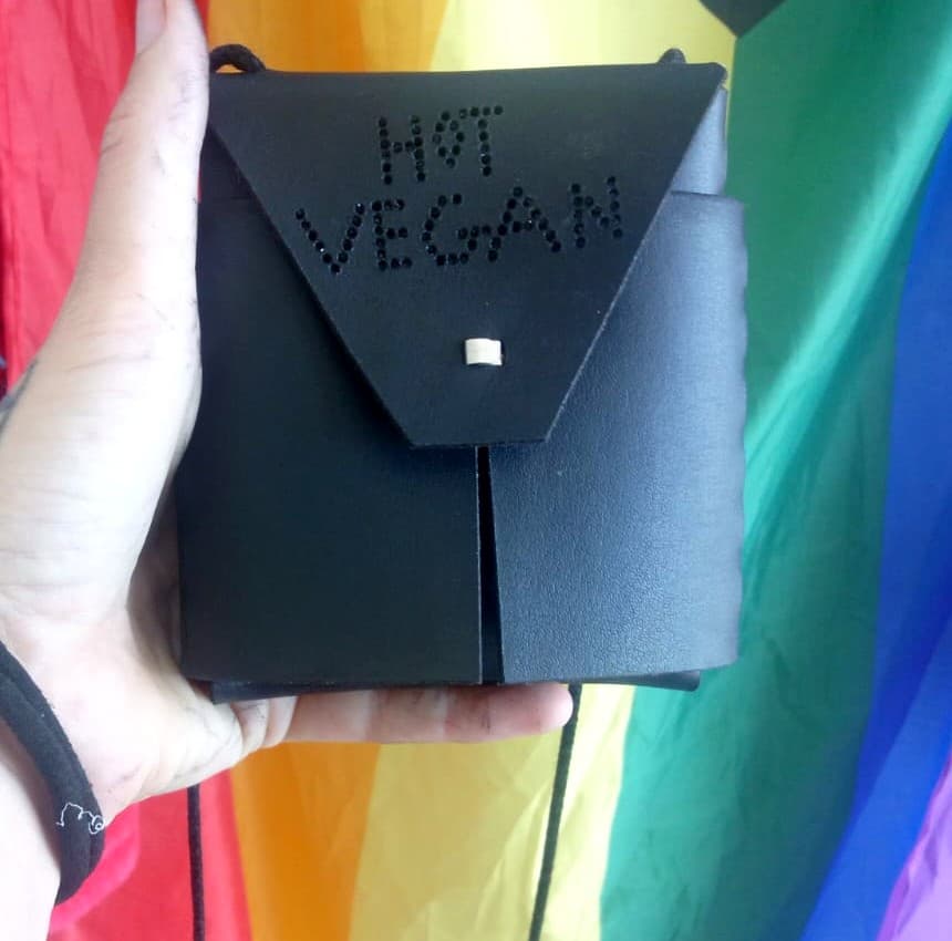 Hand holding a black cactus leather crossbody wallet with the words Hot Vegan studded into the flap, in front of a rainbow flag.